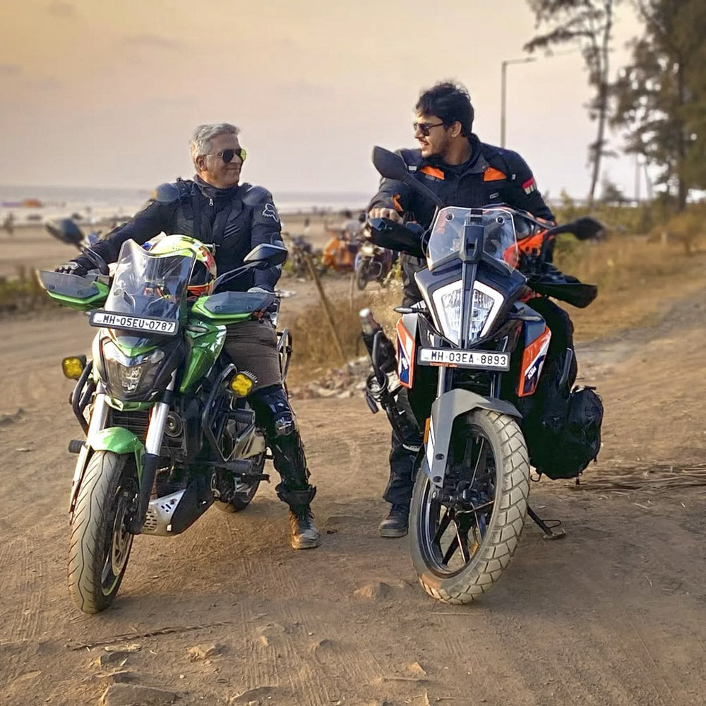 Two bikers in adventure riding gear with their motorcycles on a scenic dirt path near a beach at sunset. One rides a green touring bike, and the other a black and white adventure motorcycle. Representing the passion for motorcycling, freedom, and community—featured in the About Us section of Ink and Iron, a brand for motorcycle enthusiasts and rugged lifestyle gear.
