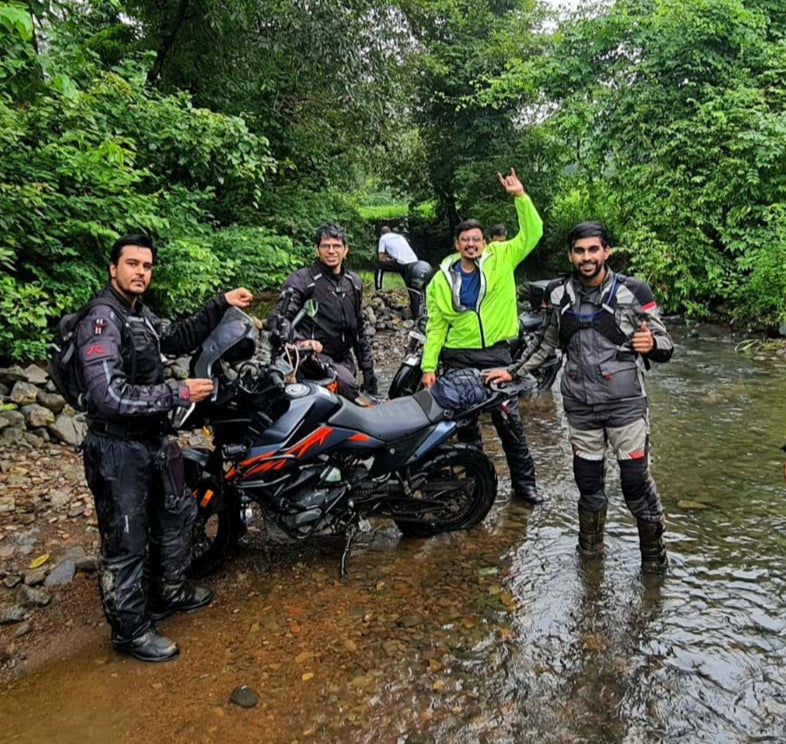 Adventure motorcycle riders crossing a water stream in lush green off-road terrain. Bikers wearing premium protective riding gear, waterproof jackets, and adventure boots while posing with their bike. Ideal motorcycle apparel for monsoon rides, touring, and off-road adventures. Explore high-quality riding gear and accessories at Ink and Iron.
