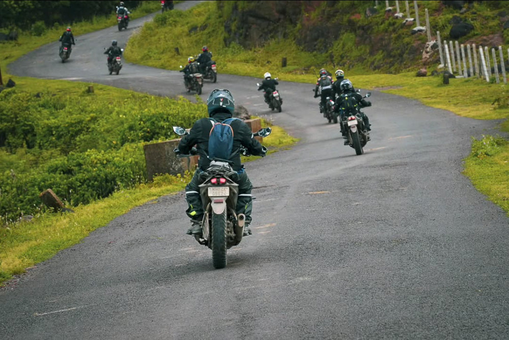 A group of motorcyclists ride along a winding mountain road surrounded by lush greenery. The lead rider, wearing a backpack and protective gear, follows the curve of the road as the group ascends into the misty hills. The overcast sky and wet pavement suggest recent rainfall, adding to the adventure's thrill.