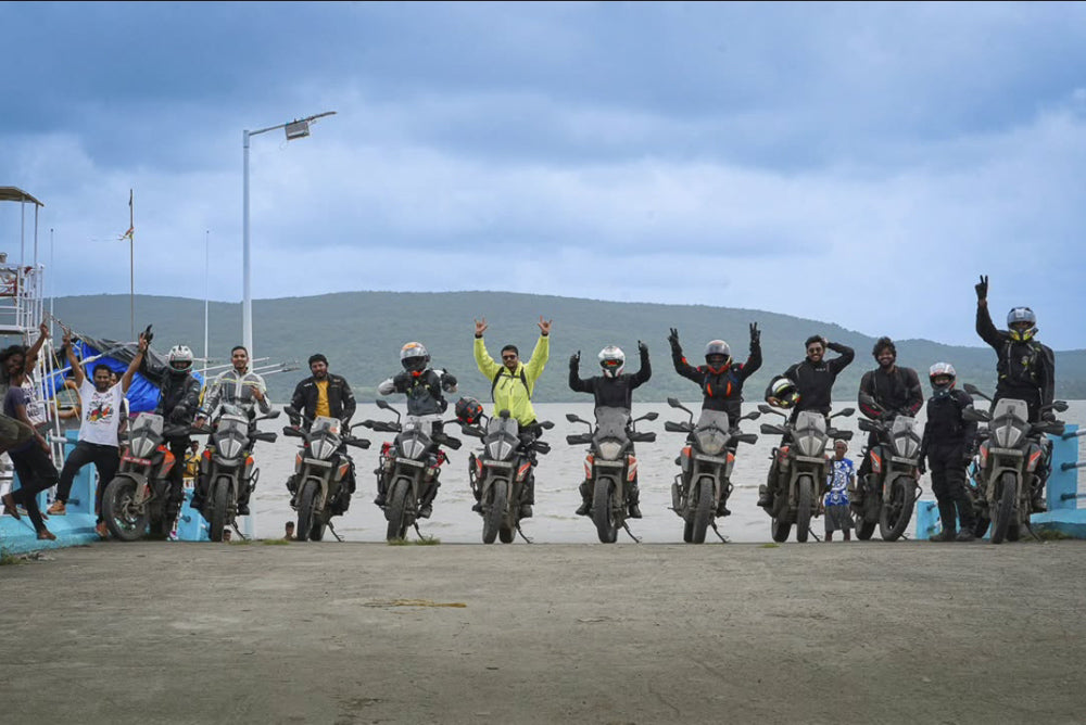 A group of adventure motorcyclists pose with their bikes on a dock, with a scenic lake and mountains in the background. Many riders have their arms raised in excitement, while others stand next to their motorcycles. The sky is overcast, adding to the dramatic setting of the journey.